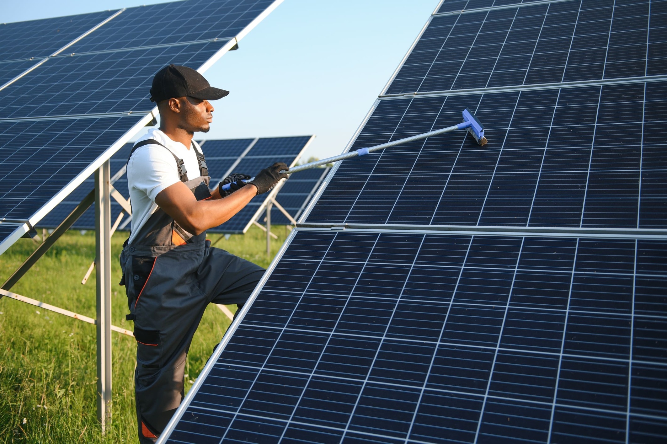 Hombre limpiando el cristal de un panel solar. Está en mitad de un prado, con paneles solares a su alrededor y de fondo.