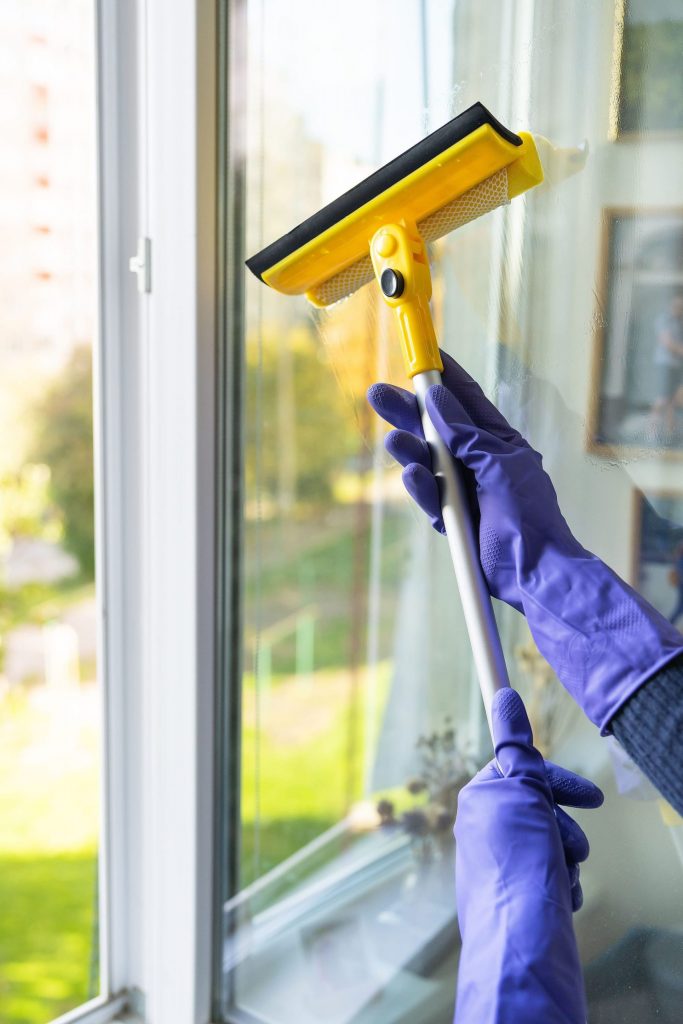 Persona usando un limpiacristales amarillo en una ventana. Se ven los brazos de la persona que lleva guantes azules. De fondo, tras el cristal, se ve un edificio y un parque, borrosos.